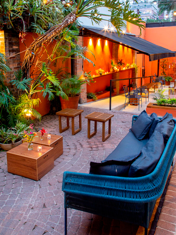 Espaço ao céu aberto, com um sofá e estofadas em destaque. Além de plantas e árvores.