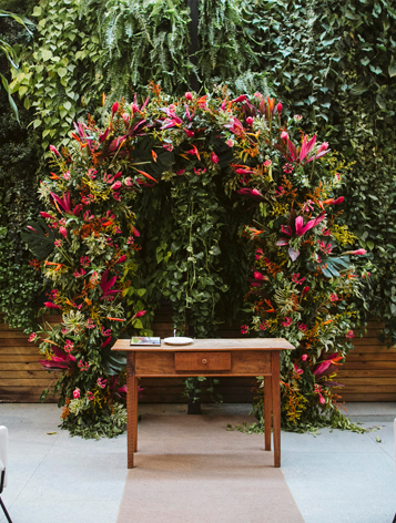 Uma mesa simples de madeira e ao fundo um arco de flores rosas com plantas verdes.