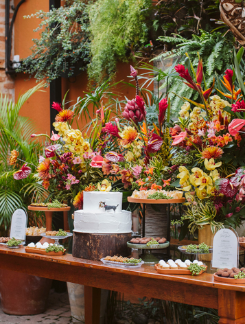 Mesa de madeira com bolos e aperitivos de casamento e flores ao fundo.