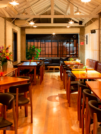 Espaço para confraternização, com várias mesas, sofá e banquetas. E iluminação ambiente e bem decorada.